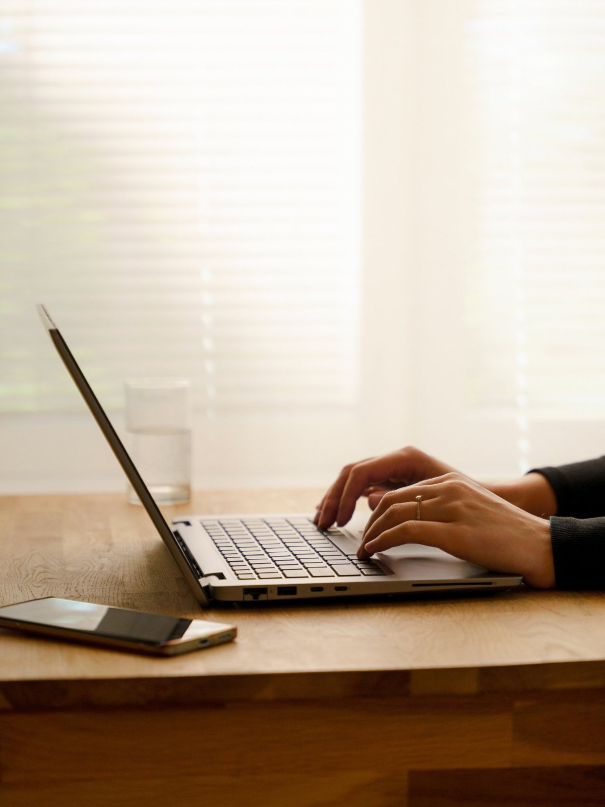 Hands typing on a laptop at a wooden desk in soft window light